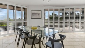 A modern dining area with a glass table and chairs, large windows with blinds, and a framed picture on the wall. - Byron Breeze 7 ~ Luxury Penthouse 3 Bedroom Apartment (Byron Bay)