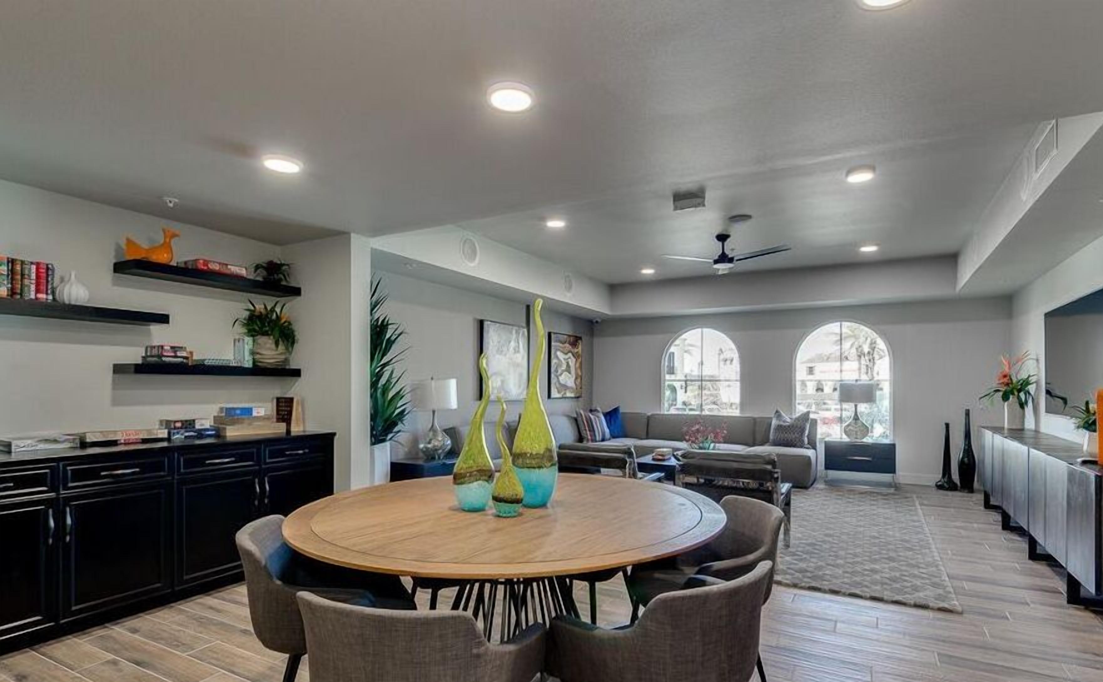 A spacious living room with a round wooden table and comfortable seating, a kitchen with dark cabinets and shelves, and large windows offering views.
