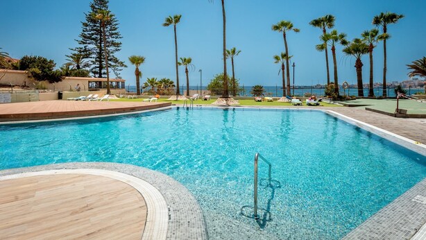Ein klares, blaues Schwimmbecken mit einer Holzterrasse, umgeben von Palmen und Blick auf das Meer.