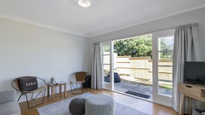 A spacious living area with a sofa, ottoman, and chairs, a wooden desk with a television, and a large sliding glass door leading to a backyard with a wooden fence. - Bach on Pitau - Mt Maunganui Holiday Home (Mt Maunganui)