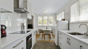 A modern kitchen with white cabinetry, a built-in oven, a gas stove, a kettle, a dining table with chairs, and a window with blinds. - Bach on Pitau - Mt Maunganui Holiday Home (Mt Maunganui)