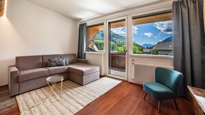Una sala de estar moderna con un sofá, una silla, una mesita y vistas a un paisaje montañoso a través de grandes ventanales.
