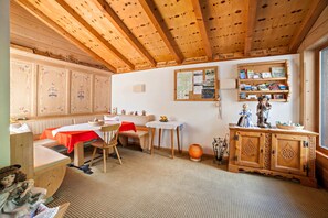 Une pièce chaleureuse avec des poutres apparentes, une table à manger, un banc, une petite table sur laquelle est posé un vase, et un meuble contenant des objets décoratifs.