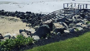 Une plage avec un escalier en bois menant à une jetée, entourée de rochers et d’une zone herbeuse.