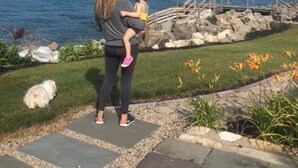 Une femme tenant un enfant près d’un quai en bois, au bord de l’eau, avec une zone herbeuse parsemée de fleurs.