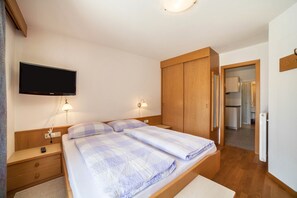 A bedroom with a bed, nightstands, a TV, wooden wardrobe, and a door leading to a kitchen.