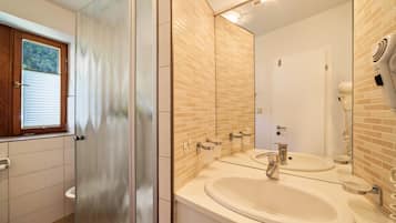A bathroom with a glass shower door, a double sink with a mirror, and a window with blinds.