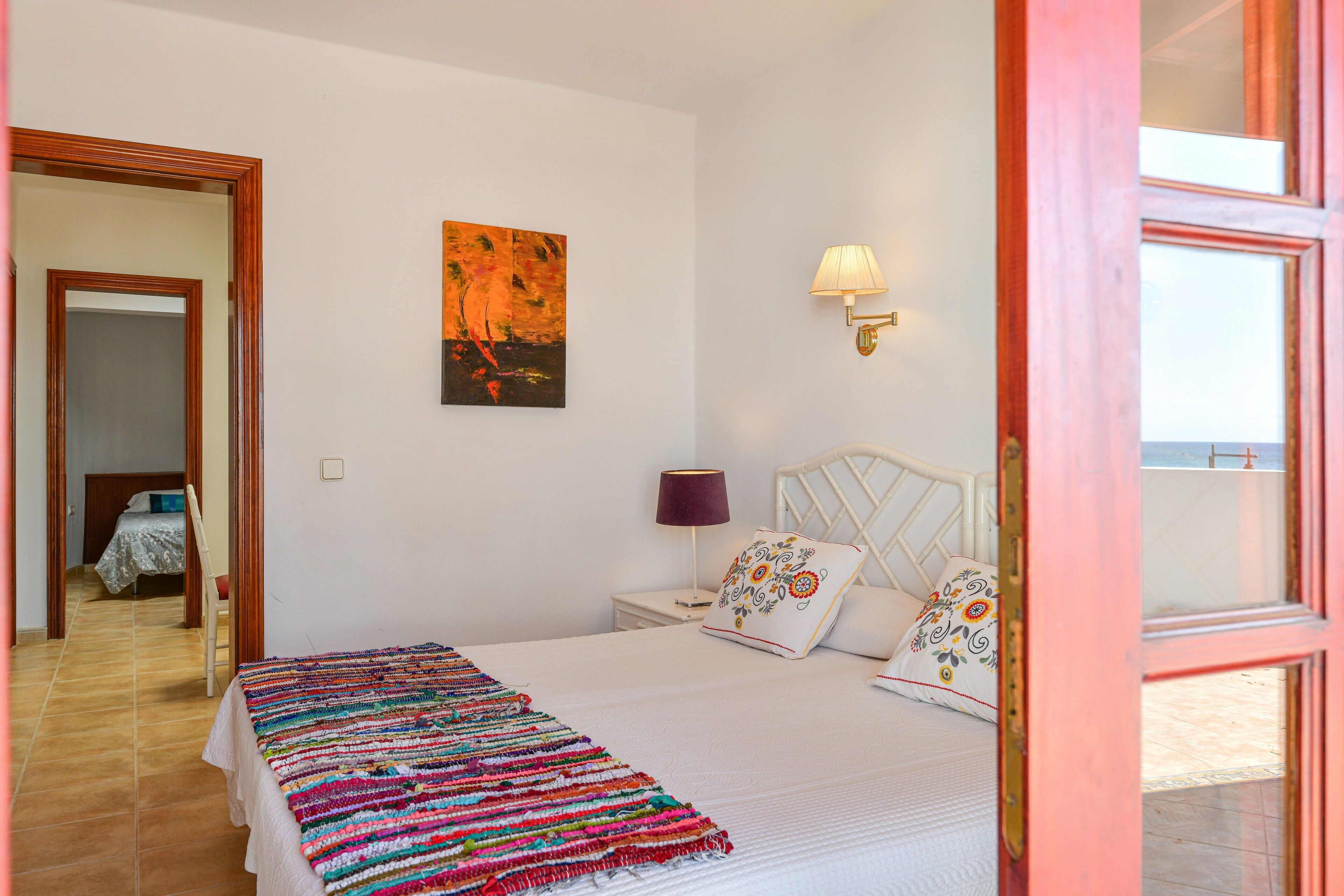 A bedroom with a bed, a nightstand, a lamp, a painting, and a view through an open door.