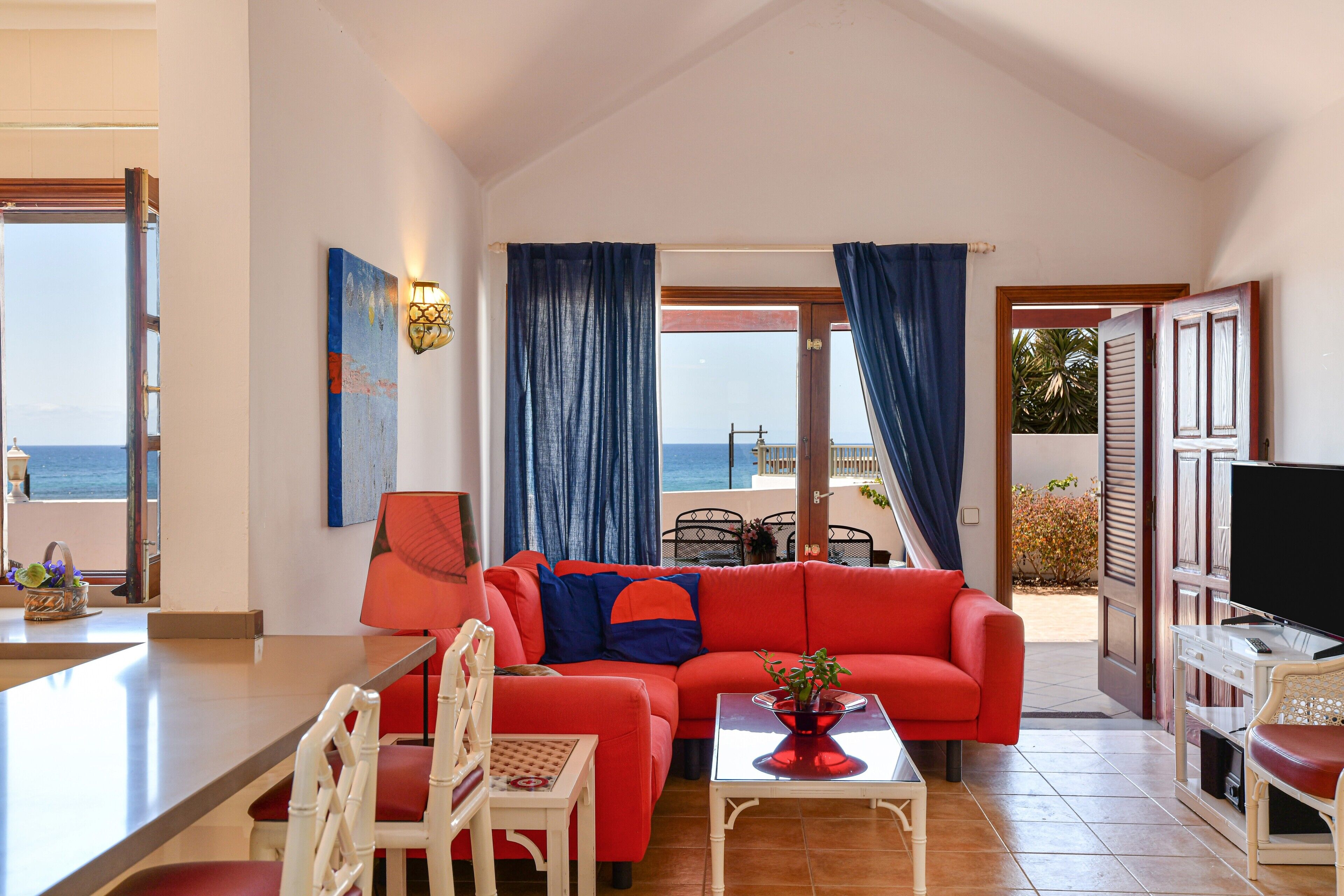 Una sala de estar con un sofá rojo, un comedor con mesa y sillas, un televisor y vista al mar a través de una puerta.
