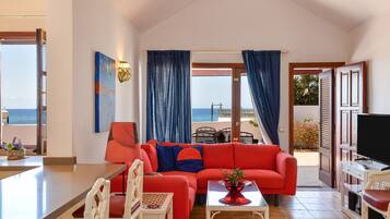 Una sala de estar con un sofá rojo, un comedor con mesa y sillas, un televisor y vista al mar a través de una puerta.