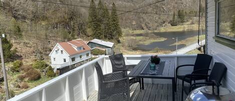 A wooden deck with a table and chairs, overlooking a valley with a house and mountains.