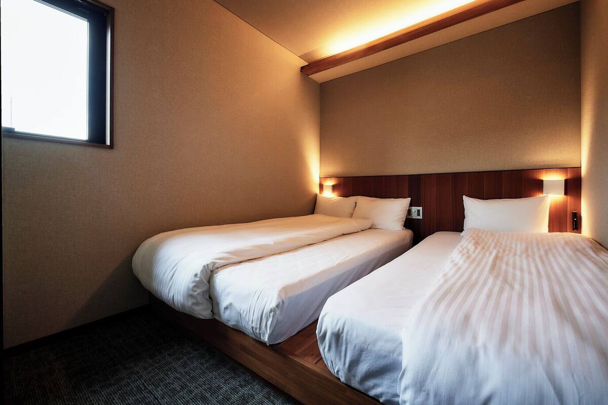 A hotel room with two single beds, a window, and a wall-mounted light fixture.