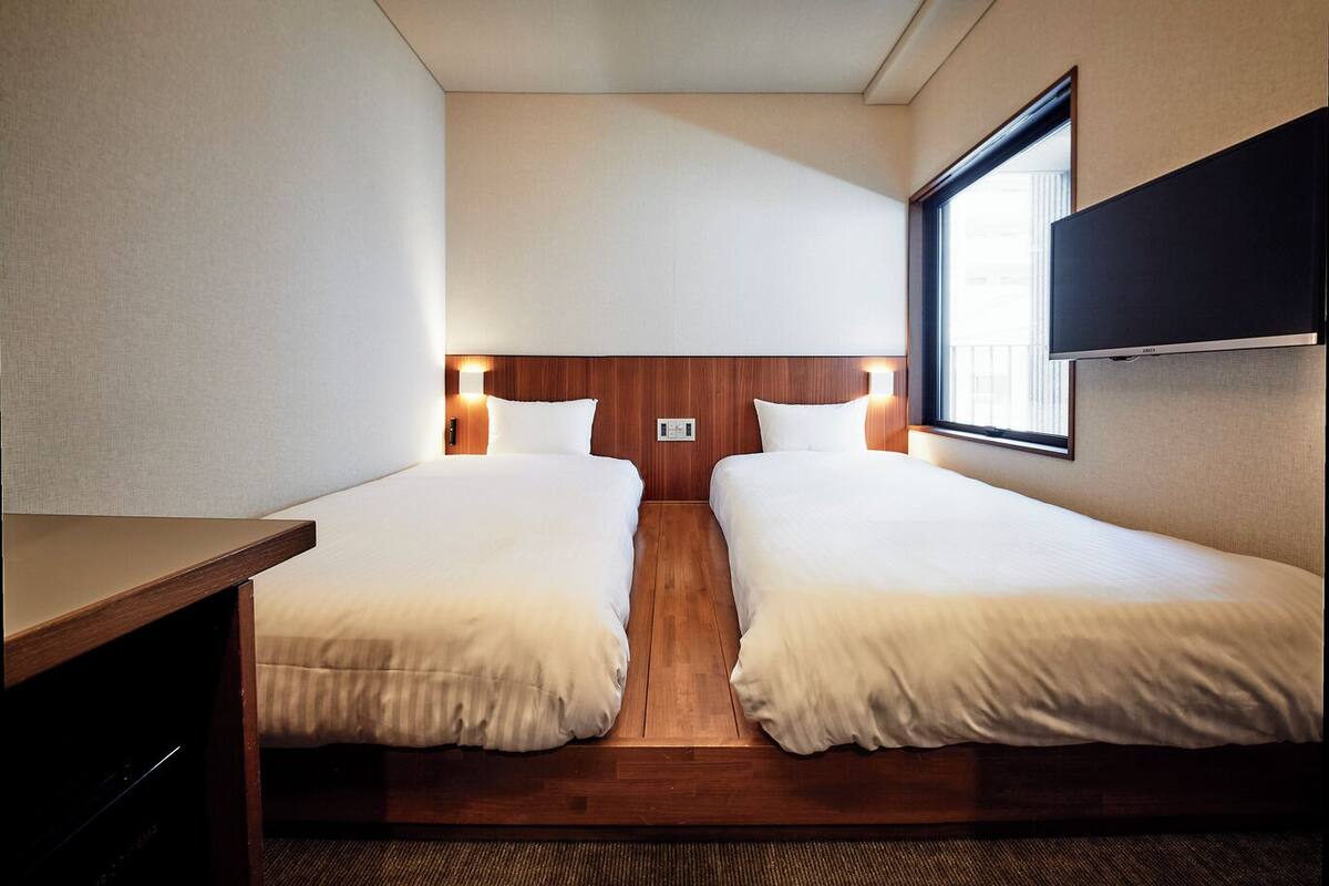 A hotel room with two single beds, a wooden headboard, a TV mounted on the wall, and a window providing natural light.