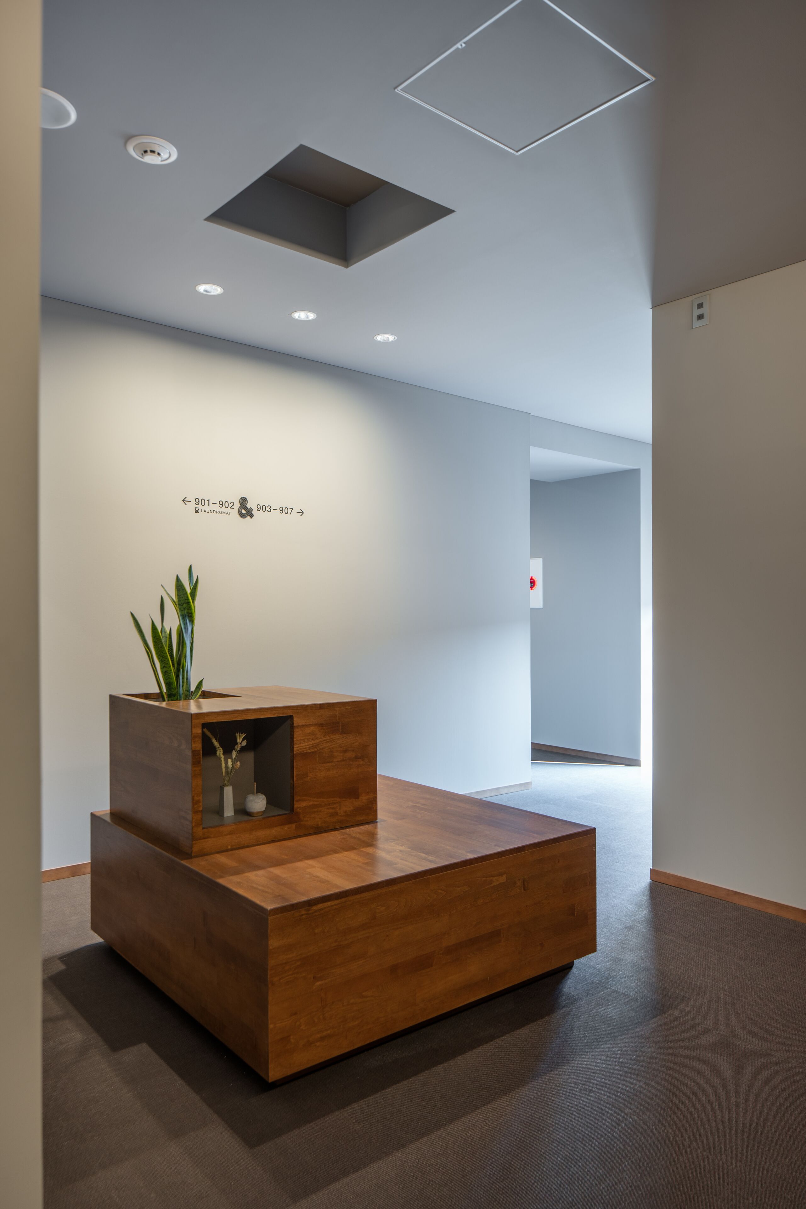 A modern reception desk with a wooden finish, a potted plant, and a wall-mounted sign with contact numbers.