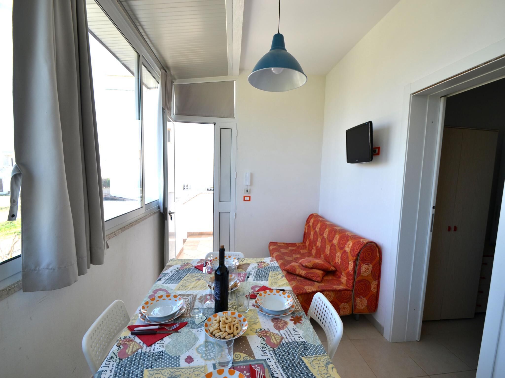 A dining area with a table set for two, a sofa, a TV mounted on the wall, and a window with curtains.