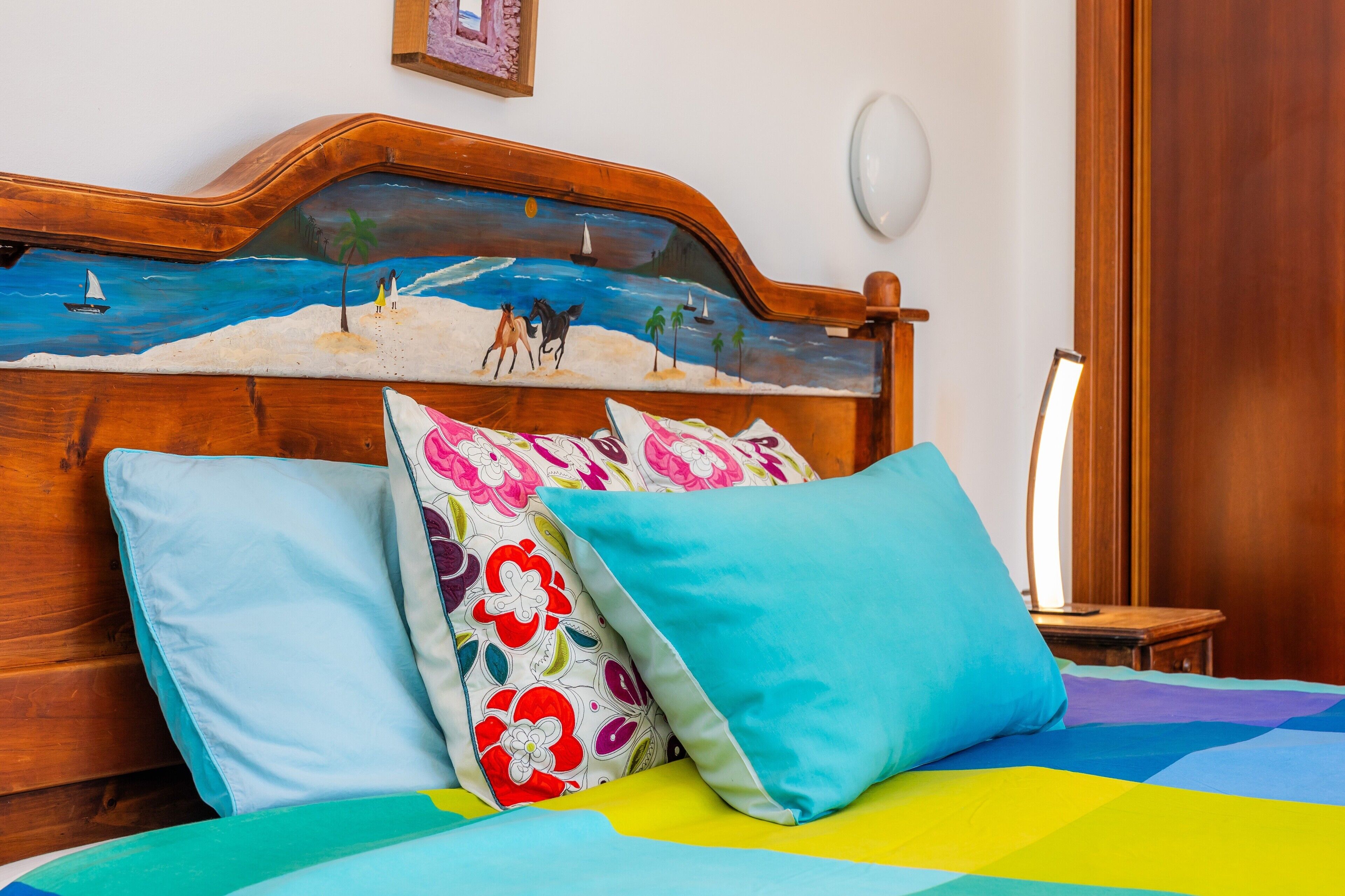 Una cama con ropa de cama y almohadas coloridas, un cabecero de madera con un dibujo pintado de playa y una mesita de noche con una lámpara.