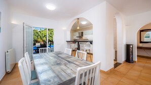A dining area with a large table, white chairs, a kitchen in the background, and a view of greenery outside. - Villa "Luna" with Sea Views, Pool, Air Conditioning, Terraces & Garden (Cala d'Or)