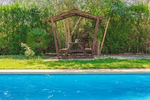 Un ensemble de balançoires en bois avec une table, installé dans un jardin luxuriant à côté d’une piscine.