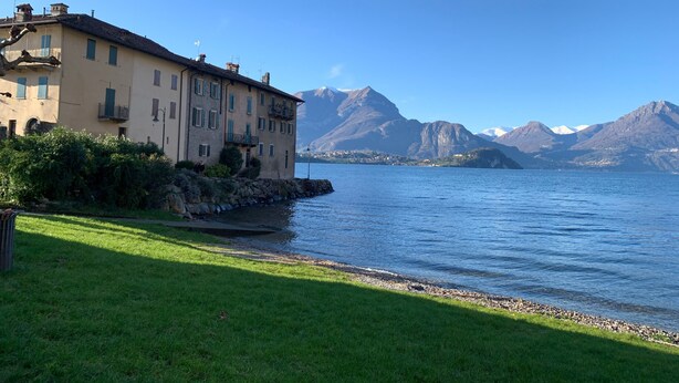 Un edificio grande con múltiples ventanas y balcones, ubicado junto a un lago con montañas al fondo.