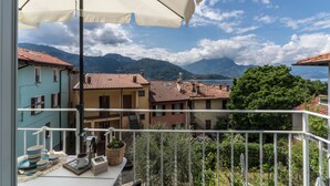 Ein Balkon mit Blick auf einen See und Berge, einem weißen Sonnenschirm sowie einem Tisch mit Geschirr und einer Pflanze.