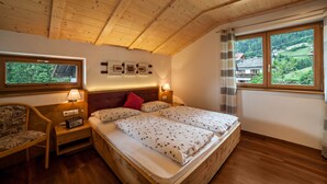 A bedroom with a wooden ceiling, a bed with floral bedding, a nightstand with a radio, a chair, and a window with a view of greenery. - Apartment "Hahndl II" with Mountain View, Wi-Fi, Garden, Terrace and Balcony (St. Leonhard in Passeier Südtirol)