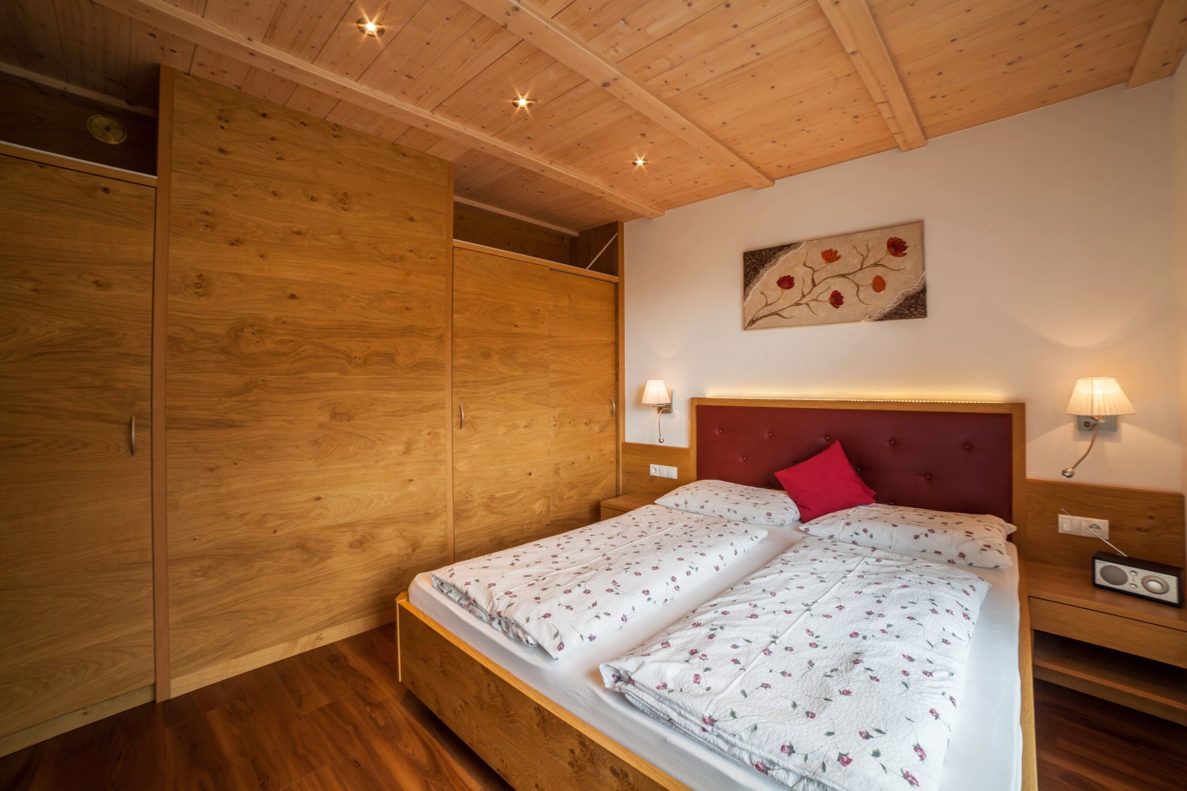 Un dormitorio con techo de madera, una cama con ropa de cama floral, una almohada roja, lámparas de noche y un radio.