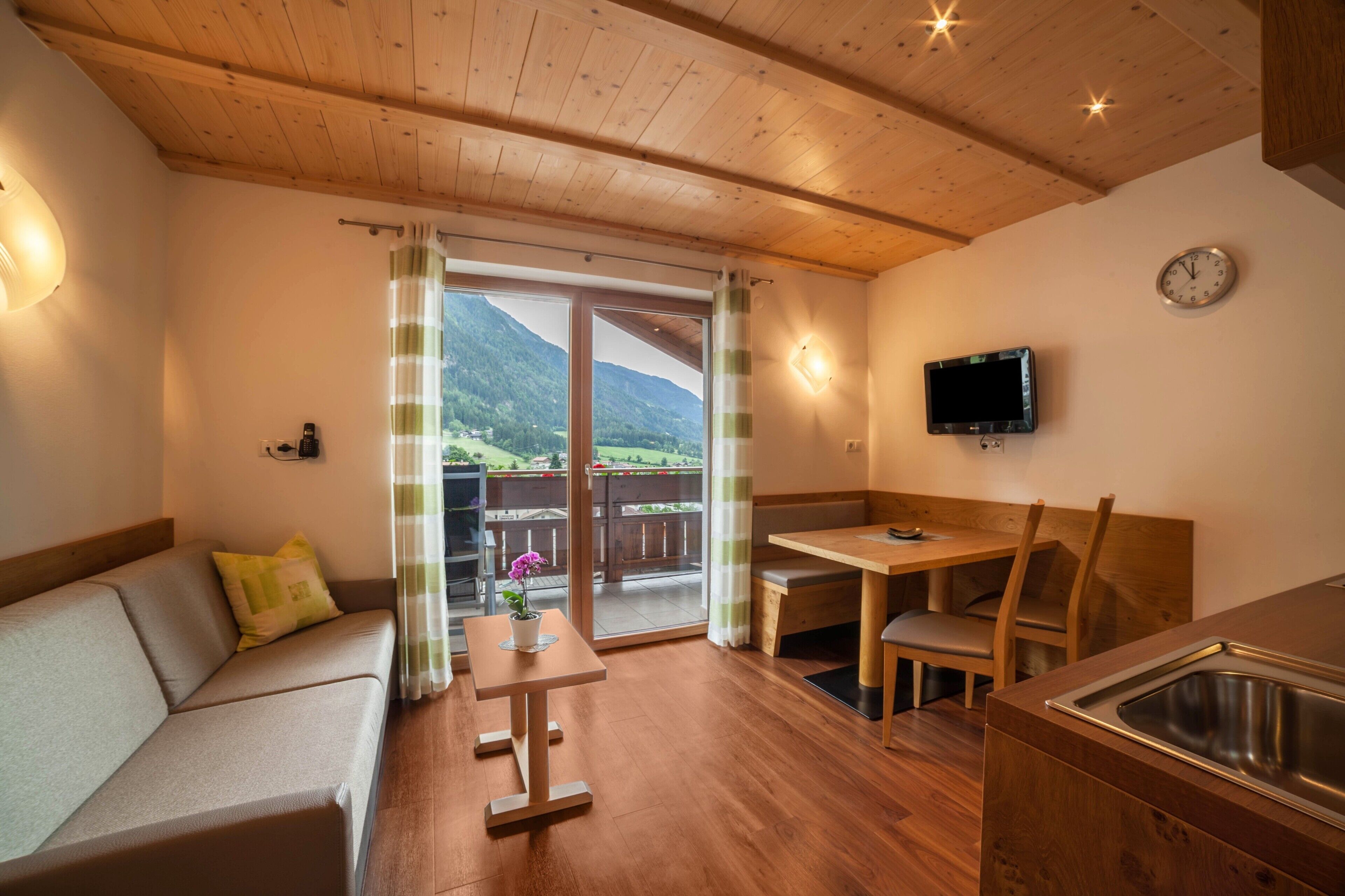 Una habitación acogedora con techo de madera, un sofá, una mesa, un banco, un televisor y vistas a las montañas a través de un ventanal.