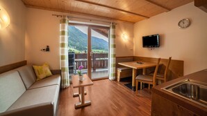 Una stanza accogliente con soffitto in legno, un divano, un tavolo, una panca, una TV e vista sulle montagne attraverso una porta in vetro.