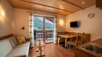 Una habitación acogedora con techo de madera, un sofá, una mesa, un banco, un televisor y vistas a las montañas a través de un ventanal.