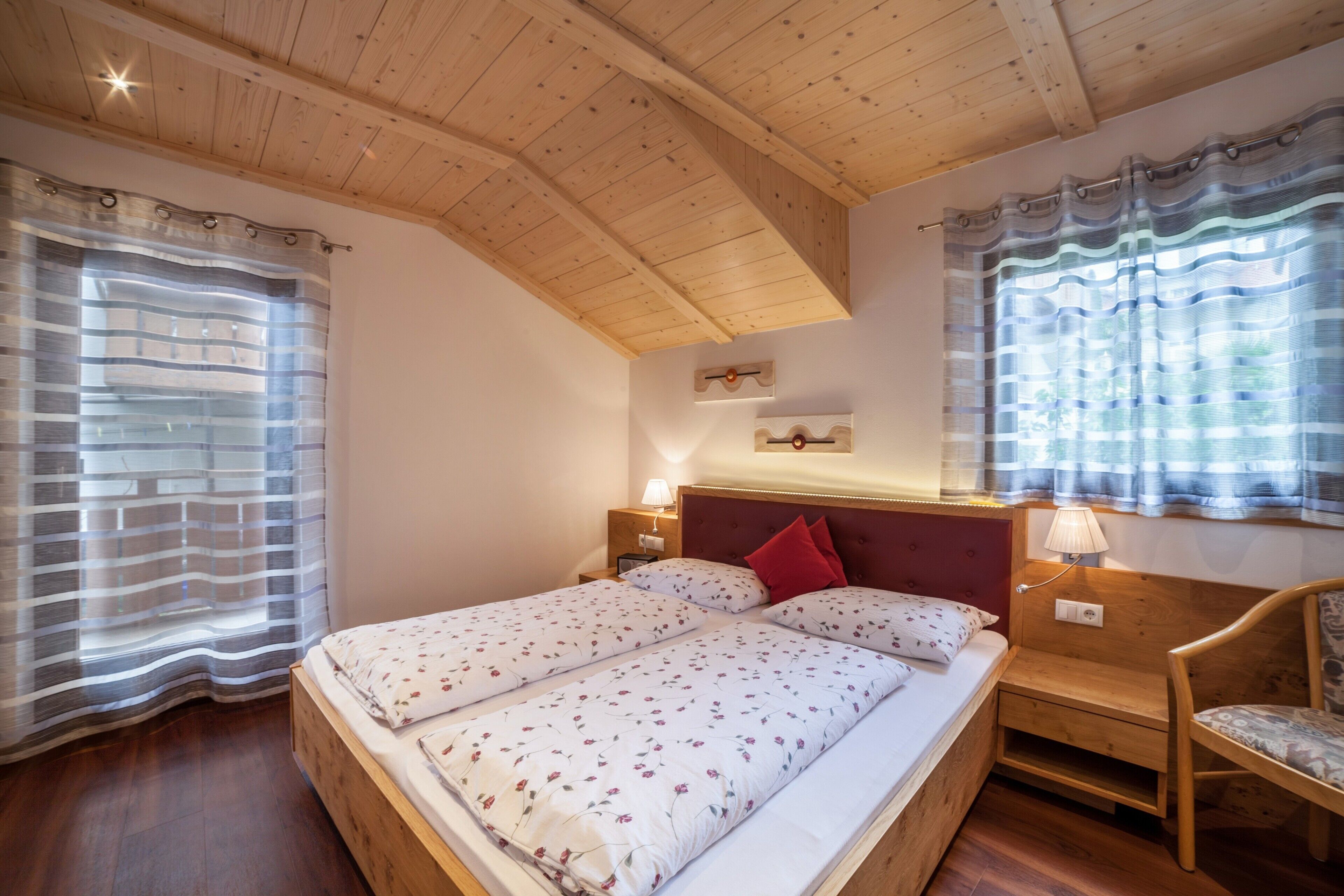 Un dormitorio con techo de madera, dos camas con ropa de cama floral, una mesita de noche con lámpara y una silla.