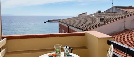 A balcony with a table set for two, overlooking a coastal view with buildings and a pier.