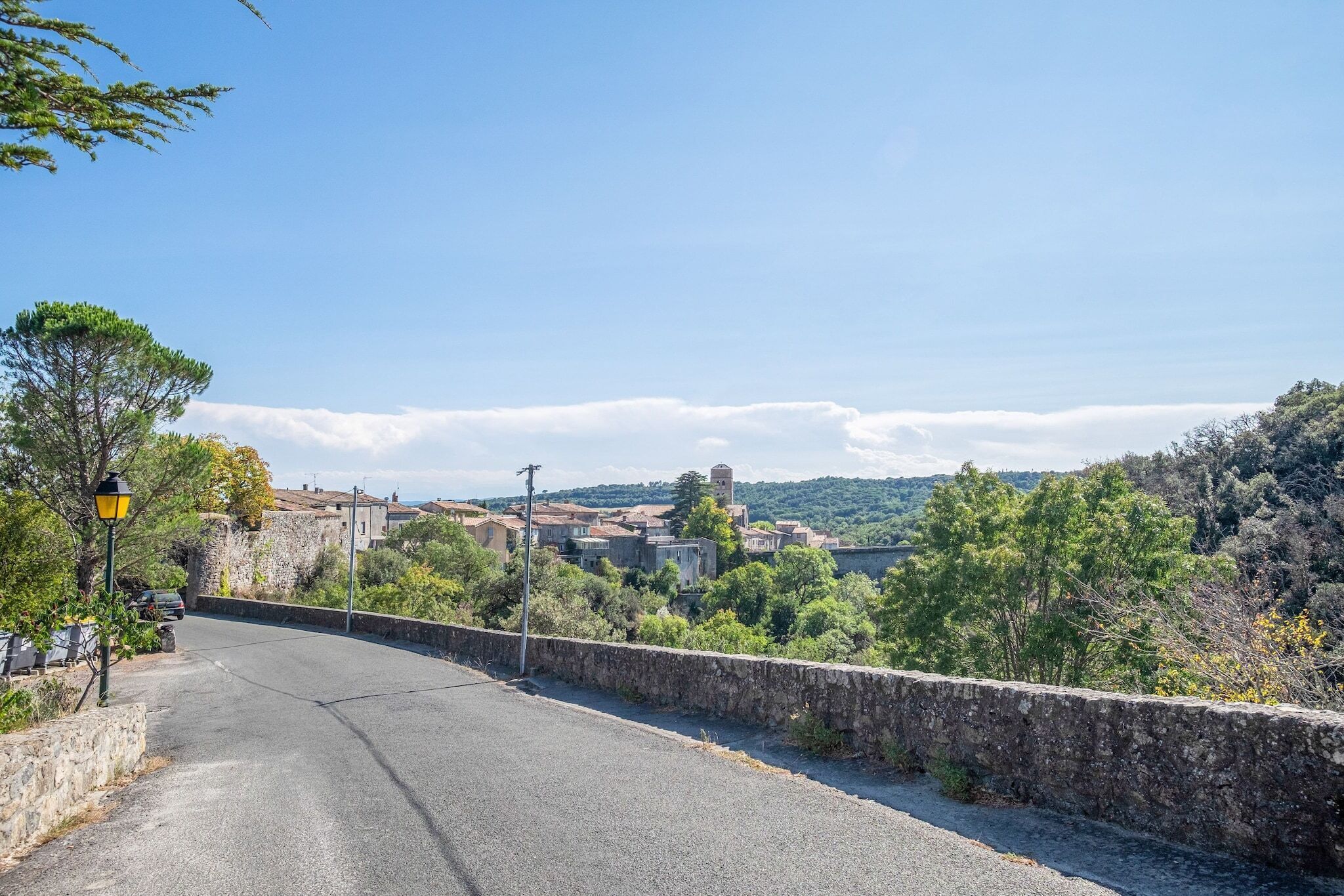 Une route goudronnée, bordée d’un côté par un mur de pierres, et qui offre une vue sur une ville avec ses bâtiments et un clocher visible au loin.