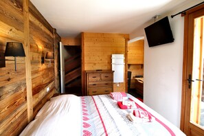 Une chambre avec un mur en bois, un lit recouvert d’une couvre-lit à motifs, une commode, un téléviseur fixé au mur et une fenêtre.