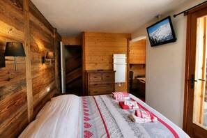 Ein Zimmer mit Holzwänden, einem Bett mit gemusterten Betttextilien, einem Schminktisch, einer Lampe und einem an der Wand befestigten Fernseher.