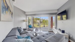 A modern living room with a grey sectional sofa, a glass coffee table, and a view of a forested landscape through large sliding glass doors. - Okanagan Living at Copper Sky #104 (West Kelowna)