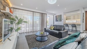 A modern living room with a stone accent wall, a round coffee table, a sofa, and a dining area with a kitchen in the background. - Modern 5B villa in El Duque, heated pool, wifi, just 250m from the beach (Costa Adeje)