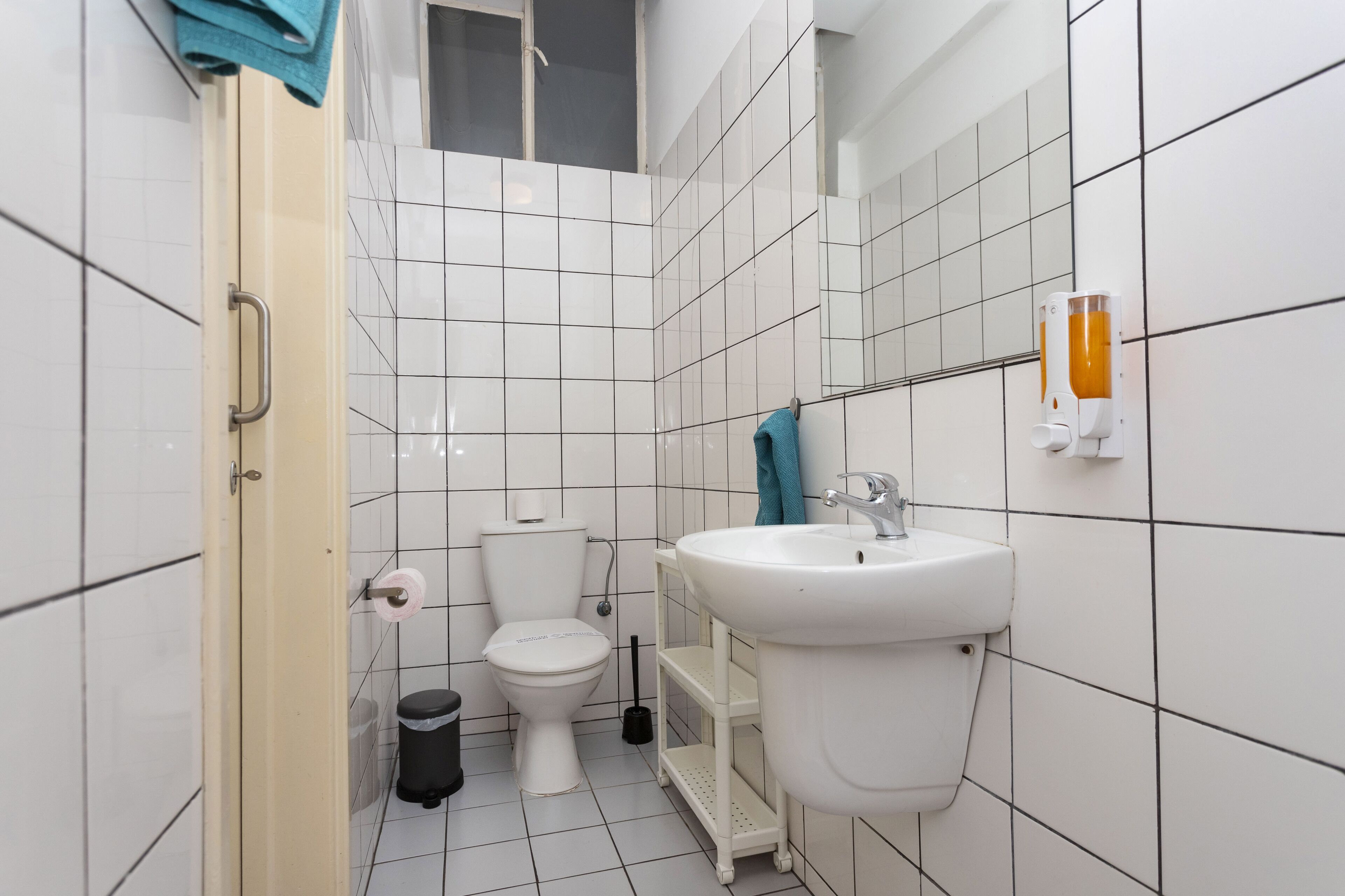 Pieni, siisti kylpyhuone, jossa on valkoiset laatat, wc-istuin, pesuallas ja käsienkuivaaja.