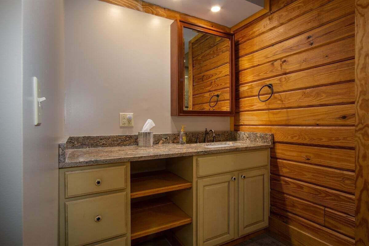 A bathroom with wooden paneling, a granite countertop, a mirror, and a towel dispenser.