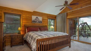 A bedroom with a wooden bed, a nightstand, a lamp, a ceiling fan, and a large window with blinds. - Blissful Bear Cabin, Long Range Views and 2 fireplaces (Brevard)