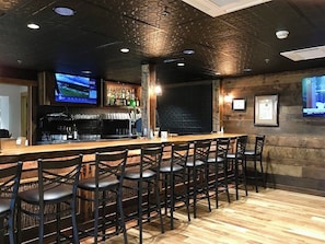 Una zona de bar con una barra larga, varios taburetes y una pantalla de televisión montada en la pared, además de una estantería con botellas.