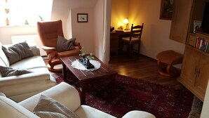 Ein Wohnzimmer mit einer Couch, einem Sessel und einem Couchtisch mit Pflanzen. Es gibt einen Essbereich mit Tisch und Stühlen sowie ein Bücherregal mit Büchern.