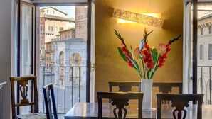 Un comedor con una mesa y sillas, un florero con flores y vistas a edificios a través de ventanas amplias.