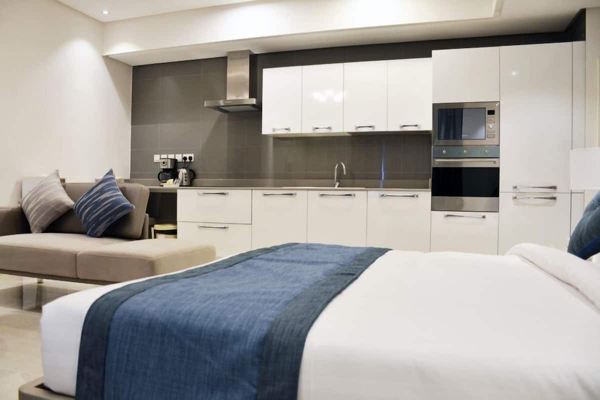 A modern hotel room with a bed, a sofa, a kitchenette, and a large window.