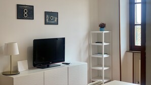 Un salon moderne avec un téléviseur à écran plat posé sur un meuble blanc, une lampe, une étagère blanche et une fenêtre donnant sur un beau paysage.