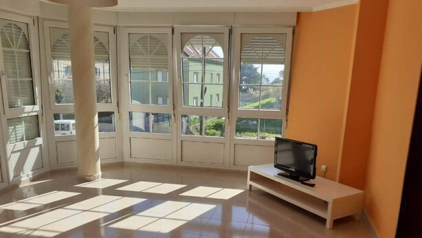 Una habitación amplia con ventanas grandes, un televisor de pantalla plana sobre un atril blanco y piso de baldosas.