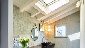 A modern bathroom with a skylight, a round mirror, a black sink, and a small stool.