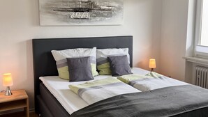 A modern bedroom with a dark-colored bed, two bedside lamps, a wooden nightstand, and a large abstract painting on the wall.