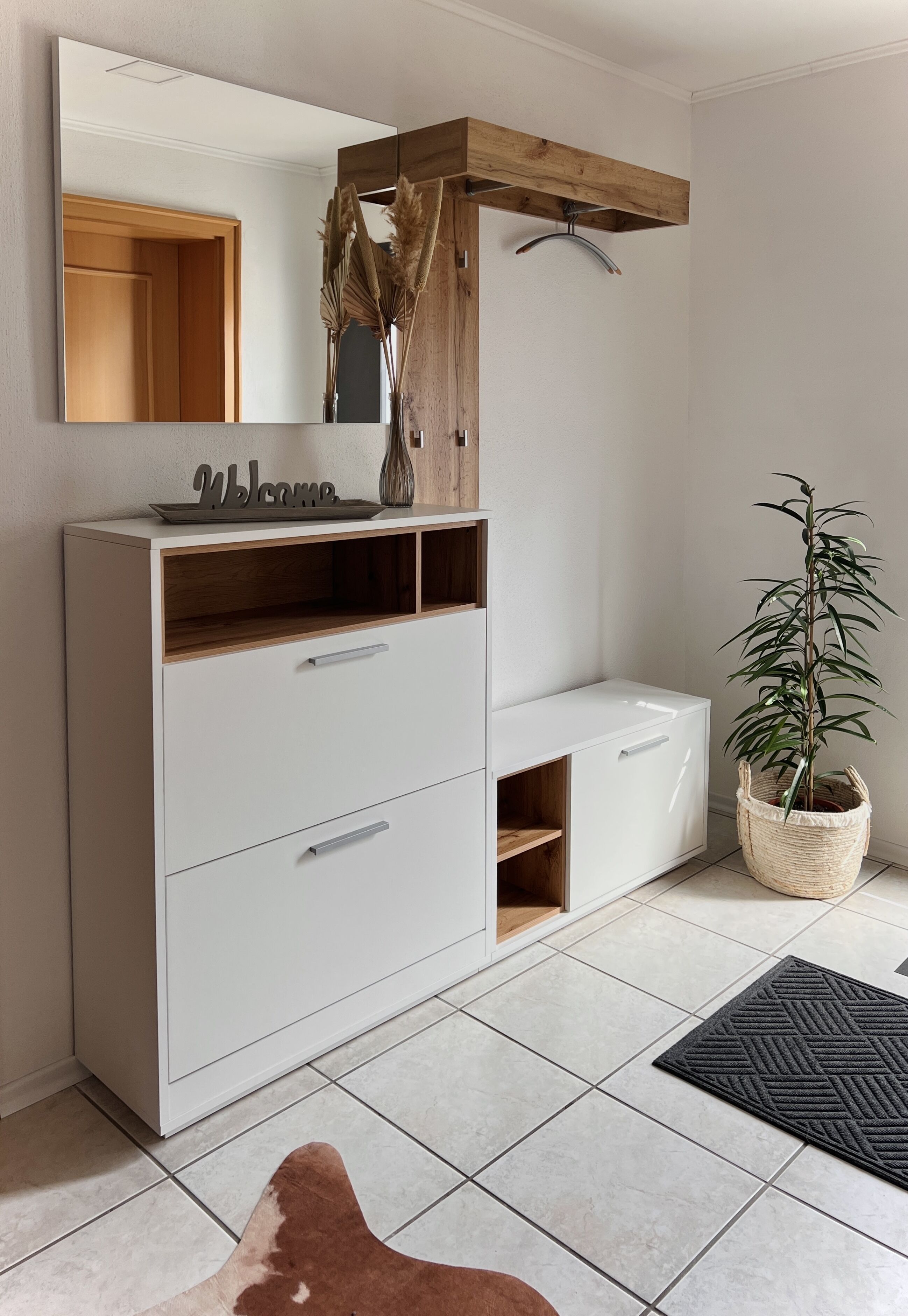 A modern bathroom with a white vanity, a wooden shelf, a hanging towel rack, a potted plant, and a brown rug.