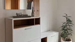 A modern bathroom with a white vanity, a wooden shelf, a hanging towel rack, a potted plant, and a brown rug.