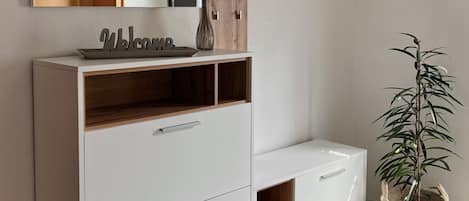 A modern bathroom with a white vanity, a wooden shelf, a hanging towel rack, a potted plant, and a brown rug.